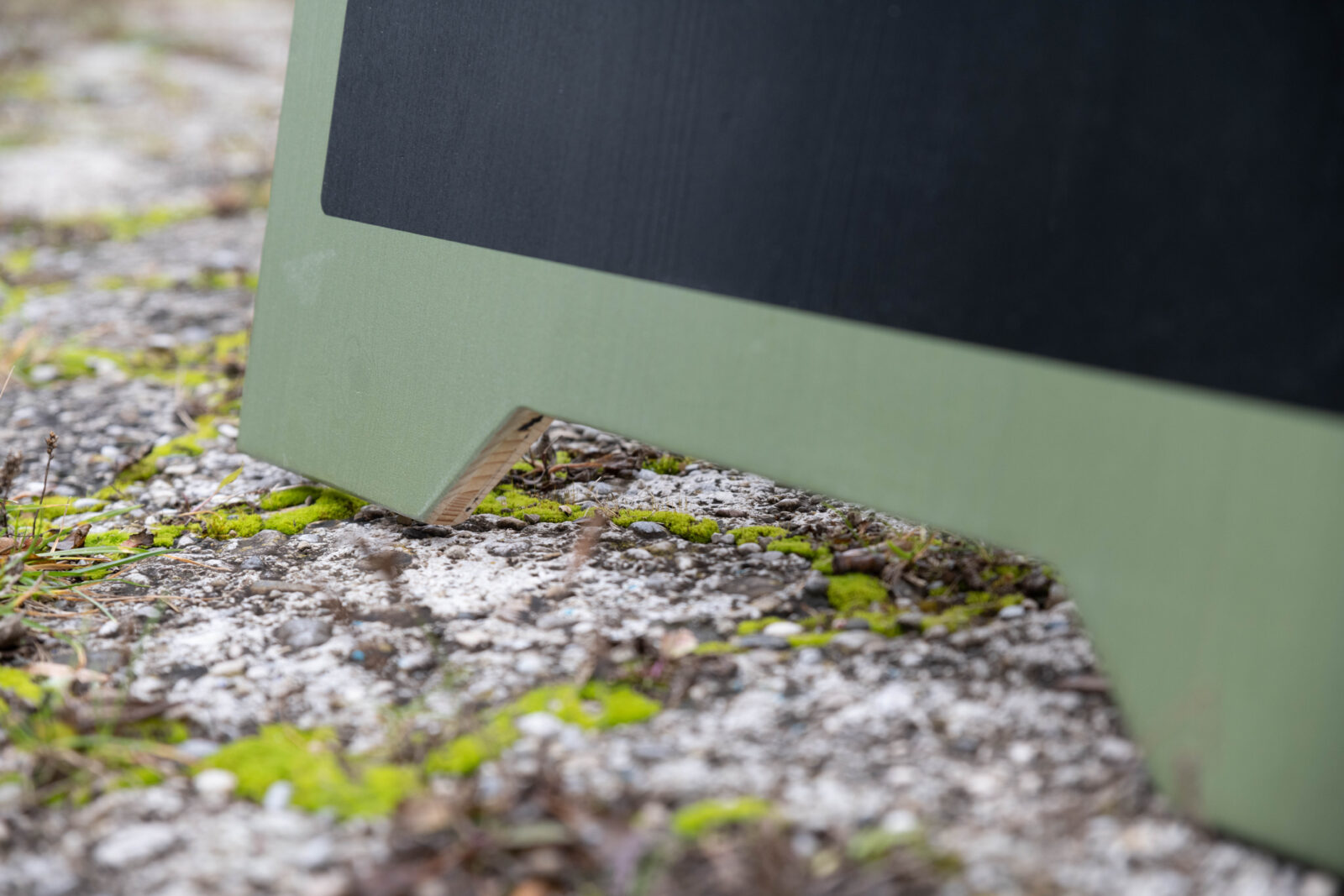 Nahaufnahme von dem Fuß eines Holz-Kundenstoppers. Man sieht den Boden mit Steine, Beton, Moos, und etwas Gras.