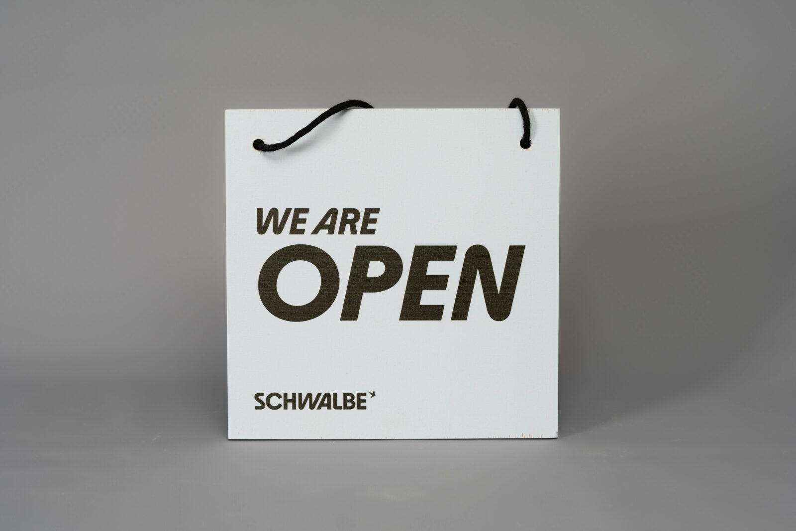 Bedrucktes Multiplex-Schild mit dem Text "WE ARE OPEN" in schwarz auf hellblauem Hintergrund, Aufhängeseil ist durch zwei Löcher gefädelt und geknotet.
